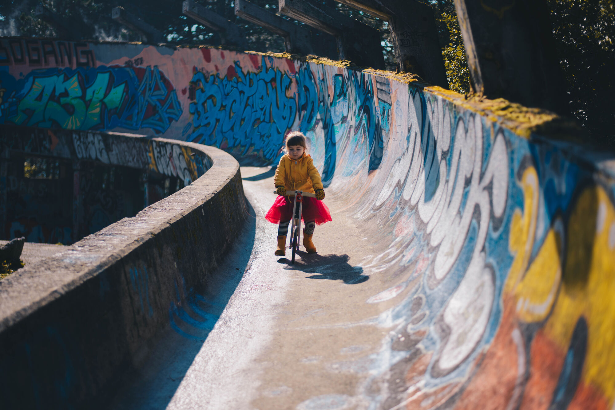 A child in a yellow raincoat and red tutu rides a wooden Tochak balance bike down the graffiti-covered concrete curve of the abandoned 1984 Olympic bobsled track on Mount Trebević.