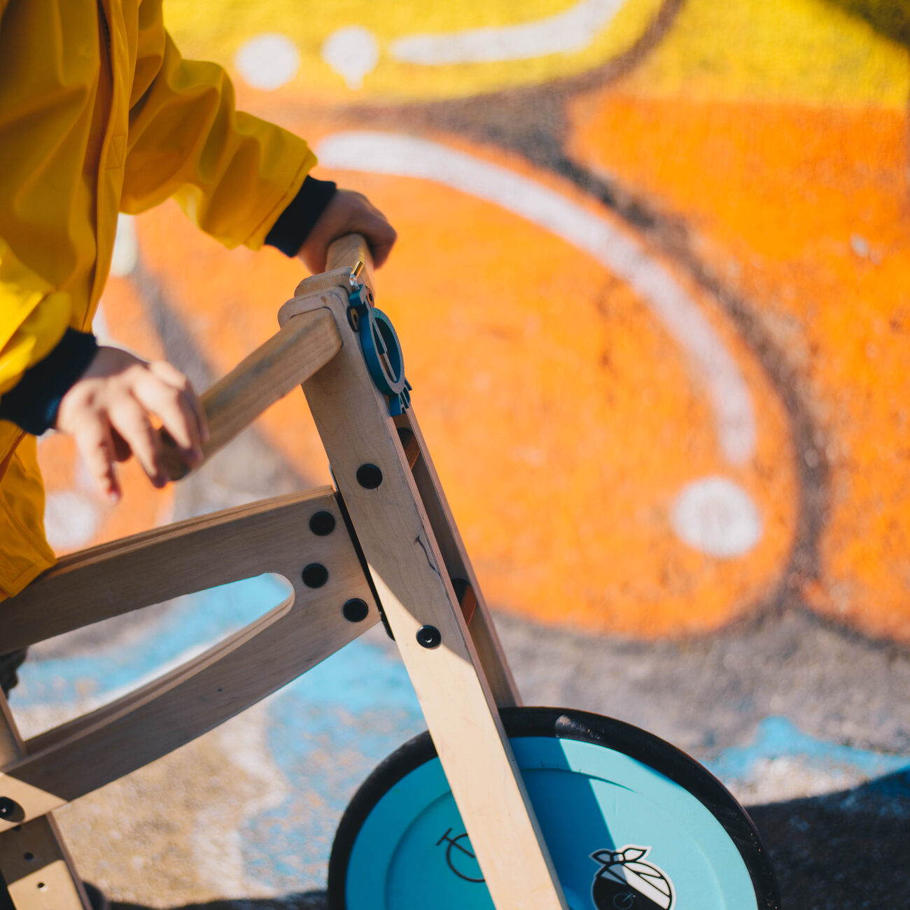 A child’s hand on the plywood handlebar of a Tochak, the blue painted front wheel and the Tochak mark below it, against orange graffiti.