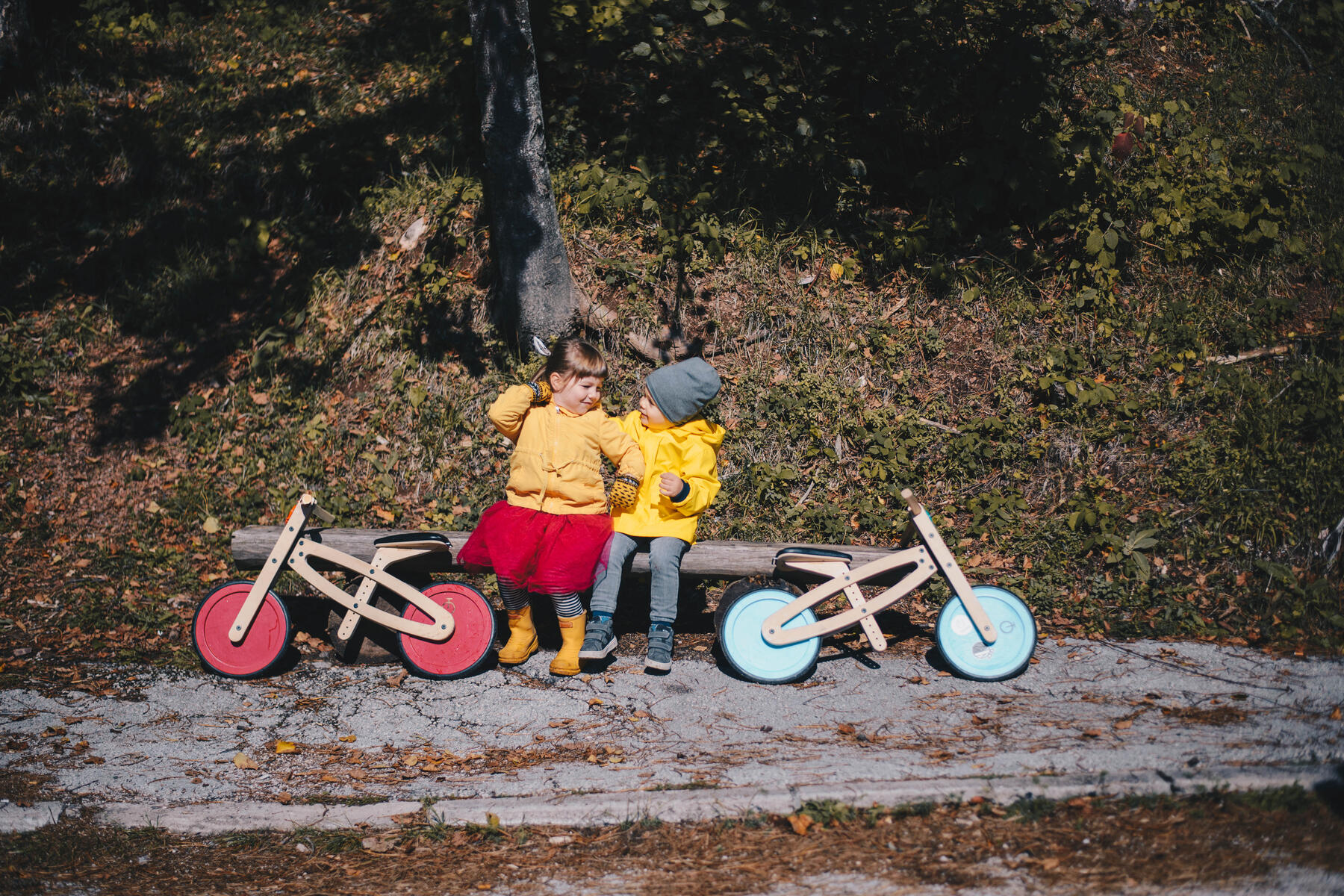 Two children sitting on a concrete ledge in the woods, a Tochak with red wheels parked on one side of them and a Tochak with blue wheels on the other.