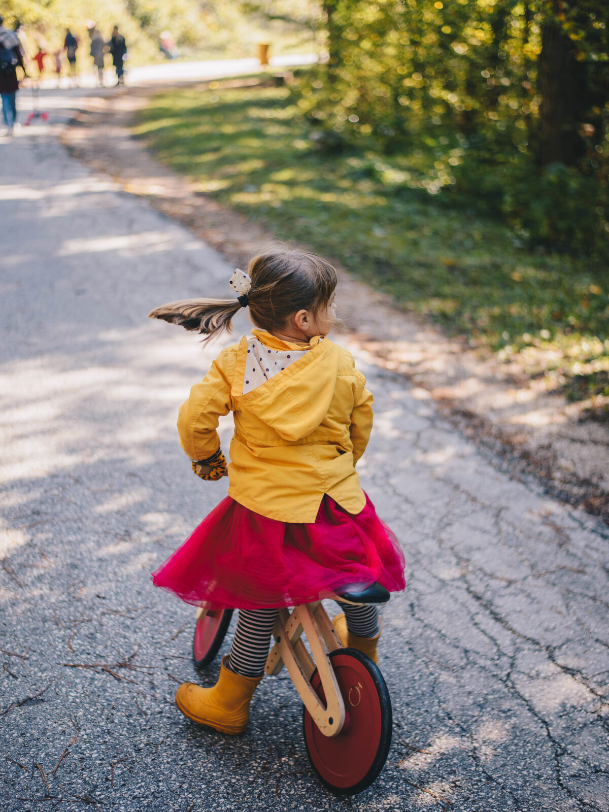 A girl in a yellow raincoat and red tutu rides away down an autumn road on a Tochak with red painted wheels.