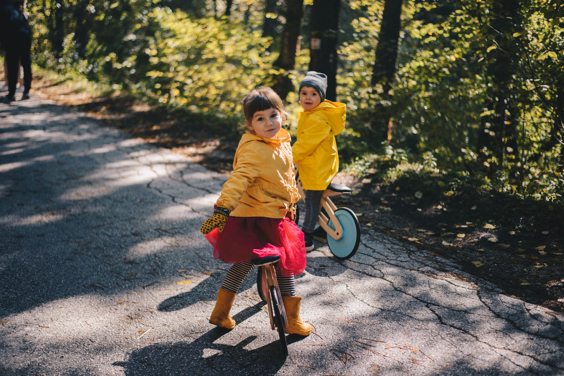 Two children in yellow raincoats on a forest road, one riding a Tochak with red painted wheels and looking back over her shoulder.